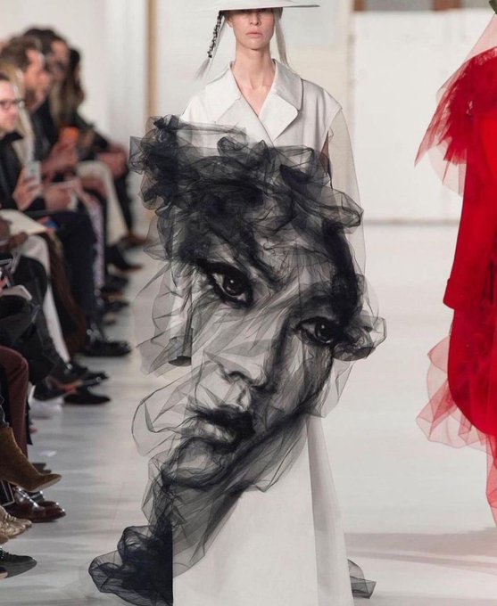 A high-fashion runway model wearing a sheer white dress featuring a dramatic black tulle overlay printed with a large face design, walking past seated spectators.
