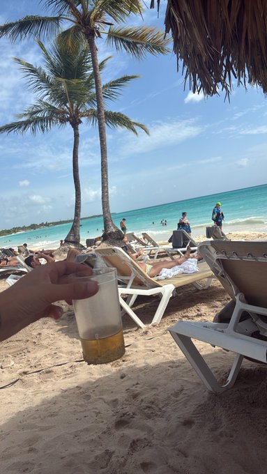 A first-person view of a hand holding a beer on a tropical beach, with palm trees, lounge chairs, and turquoise water in the background.