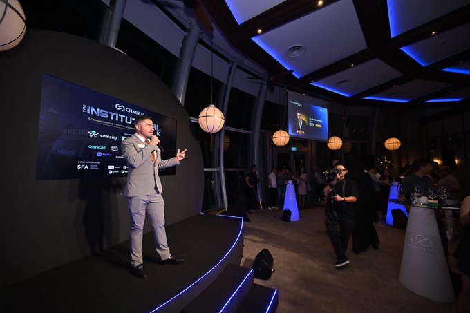 A man in a gray suit speaking into a microphone on a stage at an event, with screens behind him displaying "THE INSTITUTE" and various sponsor logos.