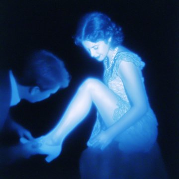 A vintage-style blue-tinted photograph showing a man in formal attire kneeling to put a shoe on a seated woman in a light, fringed flapper-style dress.