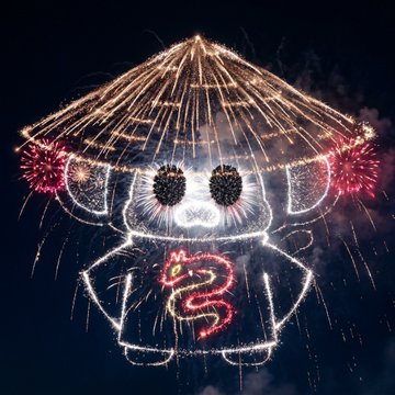 An aerial nighttime view of a drone light show forming the shape of a character wearing a conical hat, displaying red fireworks and red illuminated text.