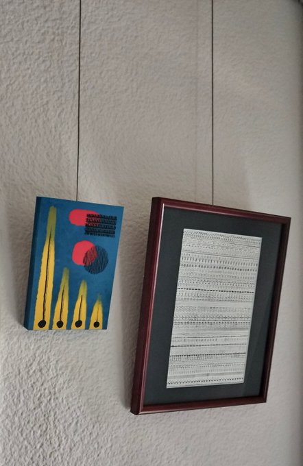 Two artworks hang on a textured white wall: a small blue canvas with abstract yellow, red, and black geometric shapes on the left, and a larger framed piece featuring rows of fine gray line patterns on the right—visually pairing an Ordinals-style digital art aesthetic with physical artwork.
