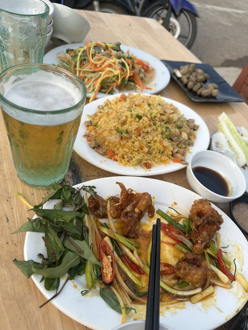 The image shows a wooden dining table with three plates of food, including rice, meat, vegetables, and noodles. The dishes are arranged neatly around the table, creating an inviting scene for a meal. A glass of beer is placed on one side of the table, adding to the relaxed atmosphere. In addition to the main dishes, there are also some bowls containing sauces or other condiments.