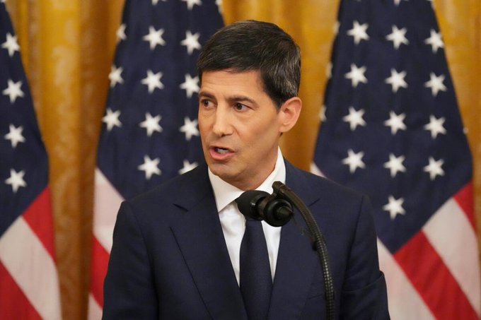 A man in a dark suit speaks at a podium with a microphone, standing before two American flags and gold drapery. The image, paired with a post about upcoming Fed Treasury bill purchases and increased liquidity, visually evokes a Federal Reserve official addressing monetary policy that could impact markets like Bitcoin.