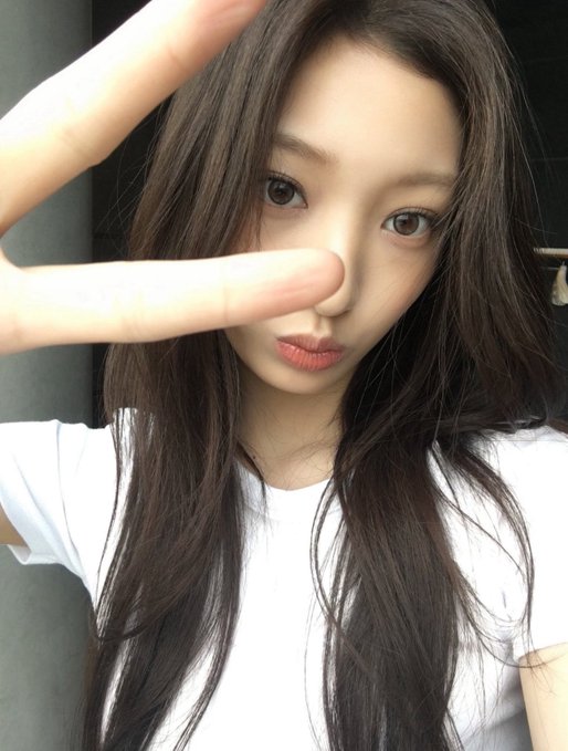 A close-up selfie of a young woman with long dark hair, making a peace sign near her face, wearing a white t-shirt and pink lipstick.