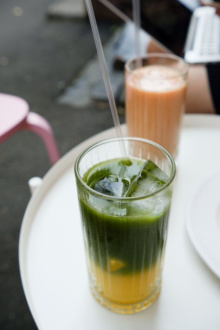 Two beverages on a white outdoor table—a layered green and yellow drink with ice in the foreground, and a pinkish-orange drink (possibly Thai milk tea) with a straw in the background.