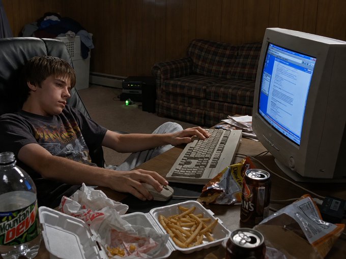 A young man sits at a cluttered desk in a wood-paneled room, using an old CRT monitor and keyboard with takeout food, a Mountain Dew bottle, and a soda can scattered around.