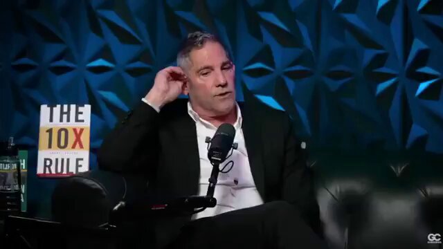 The image features a man sitting down, wearing a suit and holding a microphone, possibly giving an interview or speaking at a conference. The background of the image shows a blue wall with geometric shapes, which could be part of a stage setup for an event. There is also a bottle placed on the right side of the image.