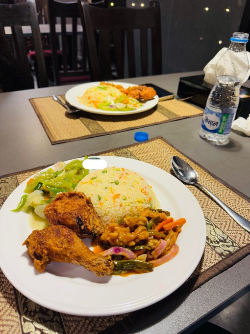 A restaurant meal on a dark table with a plate of fried chicken, fried rice, vegetables, and beans, accompanied by a bottle of Kinley water and another plate in the background.