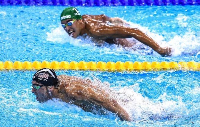 Two competitive swimmers racing in a pool, with Michael Phelps visible in the foreground lane and another swimmer wearing an arena cap in the lane behind him.