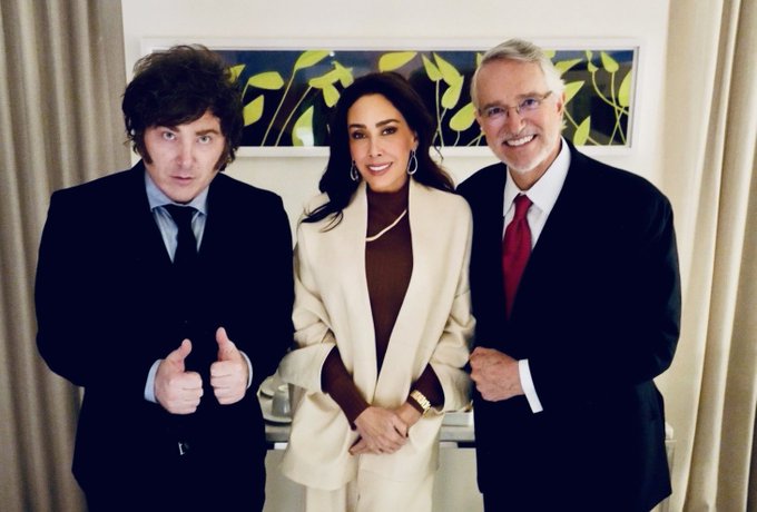 Three people pose for a photo indoors: a man on the left in a dark suit giving a thumbs-up, a woman in the center wearing a white coat over a brown top, and a man on the right in a dark suit with a red tie.