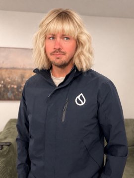 A man with shaggy blonde hair and bangs wearing a navy blue softshell jacket with a white logo stands indoors.