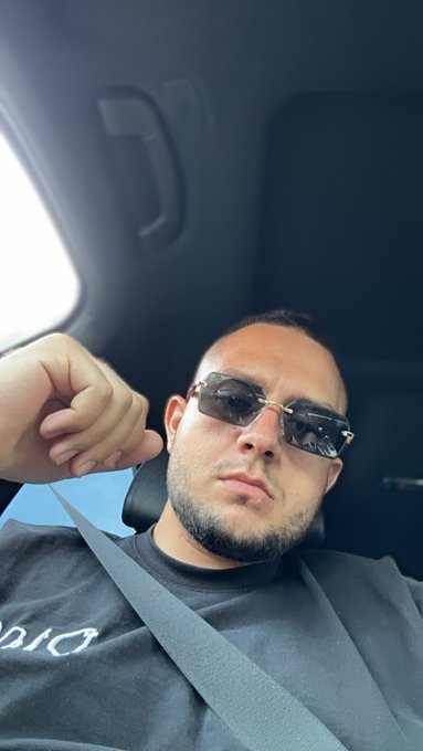 The image shows a man wearing sunglasses, sitting inside a car with his arm around another person's shoulder. The man has a beard and is dressed in a black shirt. There is no visible text or other objects in the scene that can be used to describe the image.