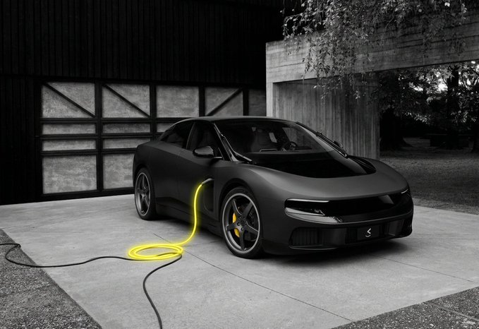 A matte black electric sedan with a glowing yellow charging cable plugged into its side is parked in front of a modern garage.