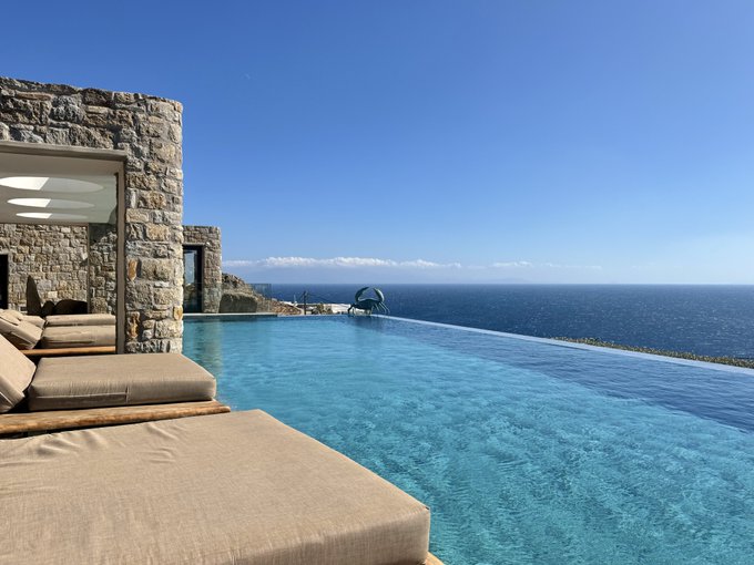 A luxurious infinity pool with beige lounge cushions overlooks a vast blue ocean under a clear sky, adjacent to a stone villa structure.
