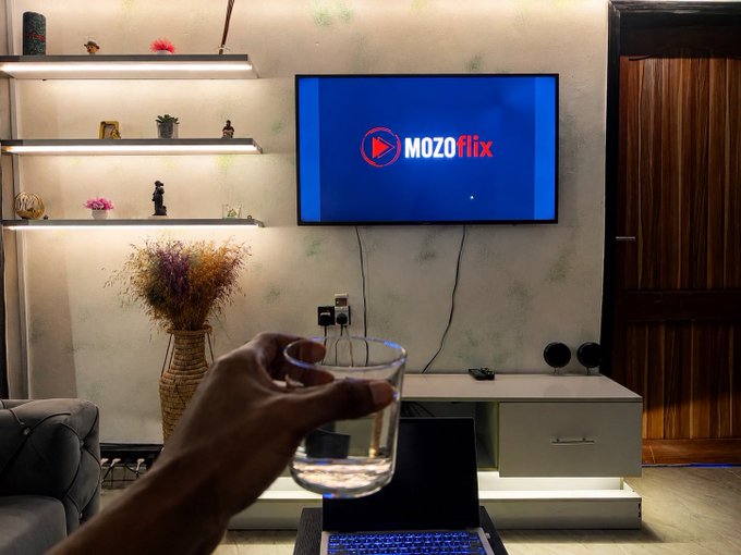 A hand holding a glass of water in a living room where a TV displays the 'MOZOflix' logo, with shelves, decorative items, and a laptop visible.
