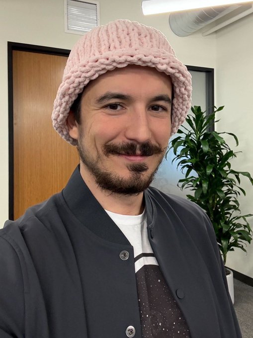 A selfie photo of a smiling man with a beard and mustache wearing a pink knitted beanie and a dark blue button-up jacket over a galaxy-patterned shirt.