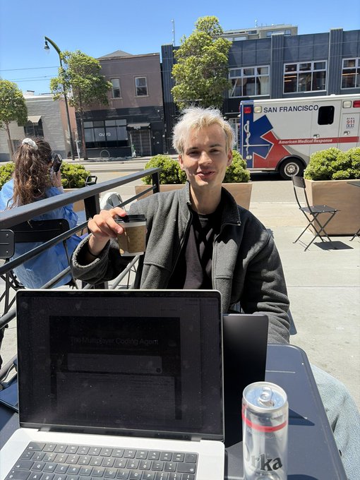 A young man with blonde hair sitting outdoors at a cafe in San Francisco (evidenced by an ambulance in the background), holding an iced coffee, with a laptop open in front of him and a red beverage can nearby.