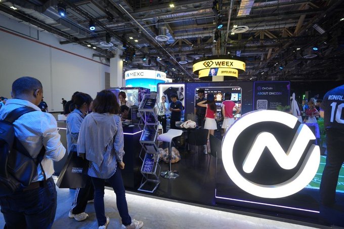 Attendees gather at the illuminated OnCoin booth at TOKEN2049, featuring a prominent lit logo display and branded materials, while neighboring WEEX signage is visible in the background.