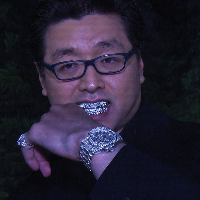 A man wearing dark-rimmed glasses and a black outfit poses with a diamond-encrusted ring in his mouth and a flashy diamond-encrusted watch on his wrist, showcasing luxury jewelry.