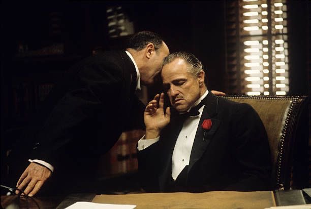 The image shows two men dressed in suits, with one man speaking into a microphone while the other listens intently. The scene takes place at an office desk or table where they are seated. In the background, there is a window that allows natural light to enter the room. A book can be seen on the desk as well. The image also contains text that reads "XRP won't be used by the DTCC". This suggests that the men might be discussing financial matters related to cryptocurrency or some other topic involving digital currency.