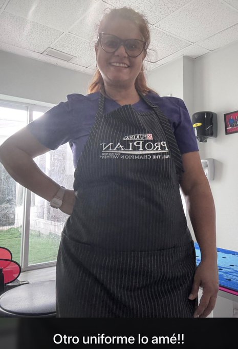 A woman with glasses and reddish hair smiles while wearing a dark striped apron over a blue shirt in a commercial kitchen setting, with Spanish text "Otro uniforme lo amé!!" at the bottom.