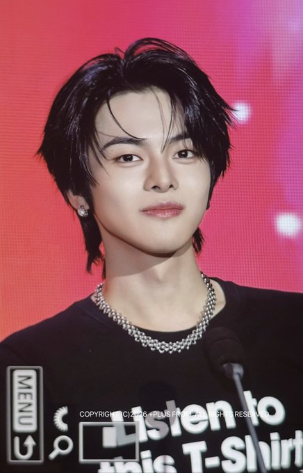 A close-up photo of a young man with dark hair wearing a silver chain necklace and black shirt against a red background, with a 'MENU' watermark.