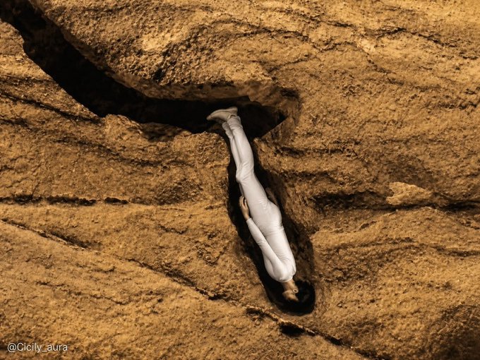 An aerial view of a person wearing a white outfit lying in the gap between large rocks, with the watermark '@Cicily_aura' in the corner.