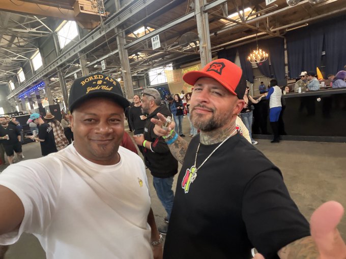 Two men posing for a selfie at what appears to be a bourbon festival; the man on the left wears a 'Bored Ape Yacht Club' cap, while the man on the right sports a red trucker hat and tattoos.
