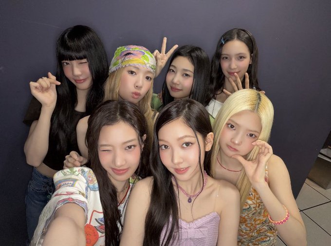 Seven young women pose together for a group selfie in front of a dark wall, making various playful gestures including peace signs and heart signs.