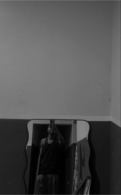 A black and white photo shows a person in a dark tank top taking a mirror selfie, with a hand partially covering their face, reflected in a uniquely shaped mirror against a plain wall.