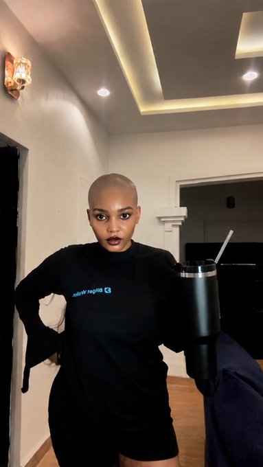 A woman with a shaved head wearing a black long-sleeved shirt and shorts, holding a large black tumbler in an indoor setting.