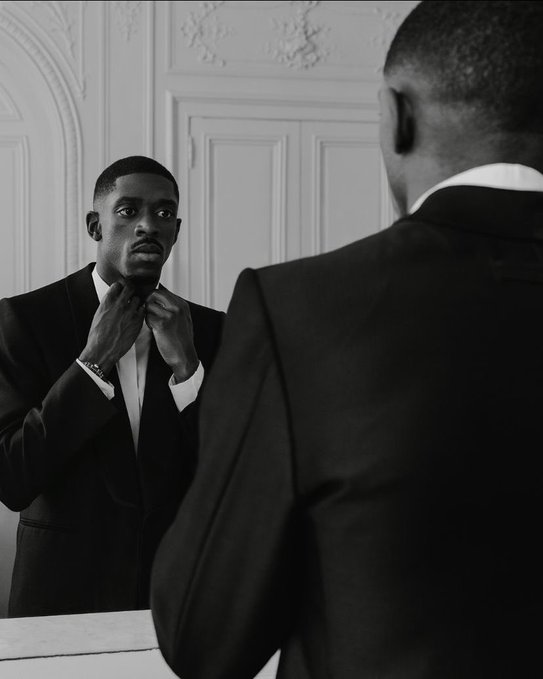 A black-and-white photo of a man in a tuxedo looking at his reflection in a mirror while adjusting his bow tie, with the back of his head visible in the foreground.