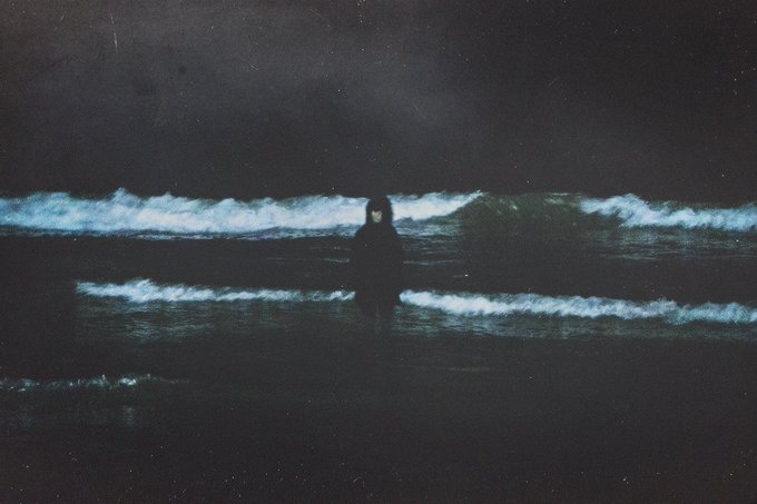 A dark, moody painting depicts a solitary figure in a black hooded coat standing in shallow ocean waves beneath a dark sky.