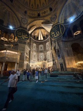 A traveler stands inside the Hagia Sophia, gazing up at the soaring domed ceiling and large circular Arabic calligraphy medallions (including the names "Allah" and "Muhammad"). The image of pausing for sightseeing reflects the post's theme of stepping away from the crypto market during a slow period to await a bull season driven by a potential Polymarket ($POLY) chain.