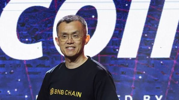 A man wearing a BNB Chain t-shirt and glasses smiles while standing in front of a conference backdrop with large blue lettering.