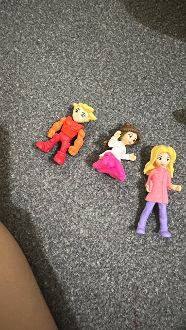 Three small handmade clay figures lying on a gray carpet, depicting cartoon-style characters with orange, pink, and blonde hair.