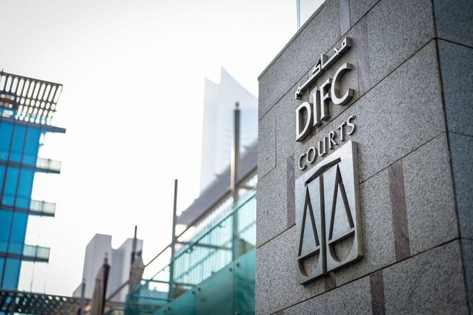A close-up of a stone sign reading 'DIFC COURTS' with a scales of justice symbol, with modern glass buildings blurred in the background, indicating the Dubai International Financial Centre Courts.