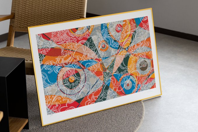 A framed, colorful artwork featuring intricate swirled patterns in red, blue, yellow, and white leans against a wicker chair on a gray rug.