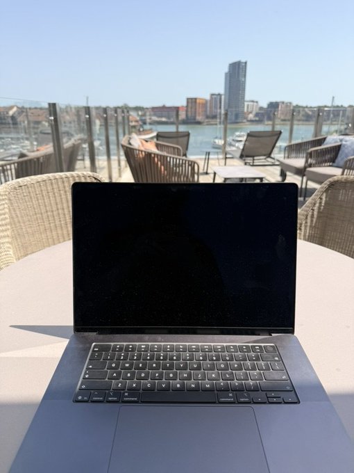 A dark laptop with a blank screen sits open on a round white outdoor table at a waterfront cafe, with a marina, boats, and city skyline visible in the sunny background.
