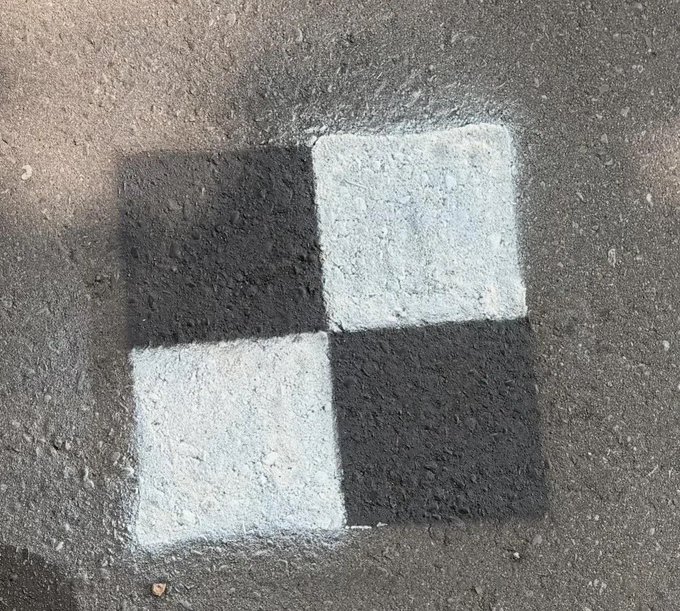 A black and white checkered square painted on asphalt pavement, resembling a minimalist street-art logo or branding symbol. The four alternating quadrants evoke a checkerboard pattern that visually echoes The Index Fund's branding style, suggesting it may be a promotional street piece for the platform.