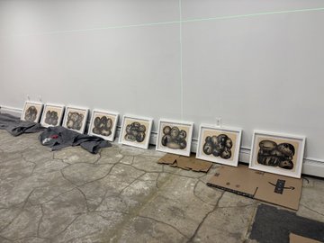 A row of framed illustrations of faces displayed on a sidewalk in front of a white wall, with cardboard laid out on the pavement.