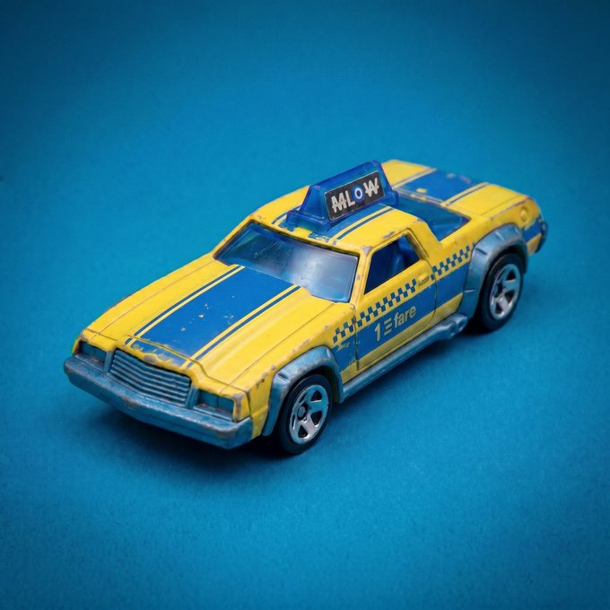 A toy taxi cab with "MLOW" on its rooftop sign and "1 fare" on its side, painted in a yellow and blue racing livery with checkered accents, photographed against a blue background. The model reflects the honorary Racer skin design celebrating NYC cab artwork, minted as an additional piece for MLow's Racer collection.