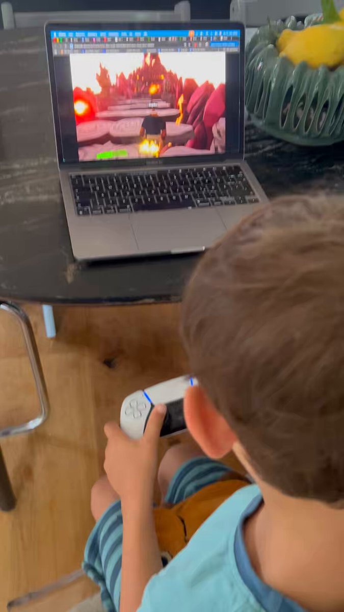 The image shows a young boy sitting at a table, playing video games with a remote controller in front of him. The laptop he's using displays an exciting game that has captured his attention. The surrounding environment includes chairs placed around the dining table where the child is seated and a bowl on the table next to it.