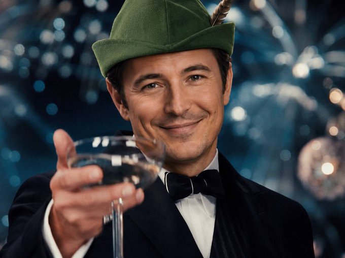 A man in formal black tie attire wears a green Robin Hood-style hat with a feather and holds up a champagne glass in a toast, smiling against a sparkly blue background.