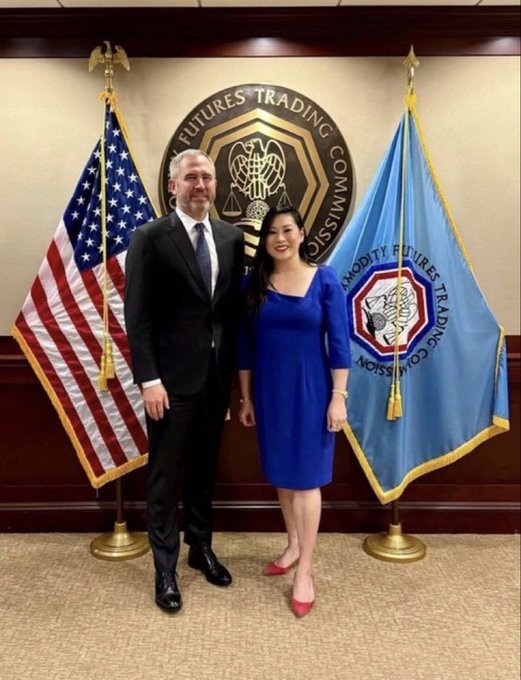 A man in a dark suit and a woman in a blue dress pose together in front of a "Futures Trading Commission" seal, flanked by an American flag and a Commodity Futures Trading Commission flag. The formal setting suggests an official meeting or announcement at the CFTC, aligning with the "BIG ANNOUNCEMENT" caption.