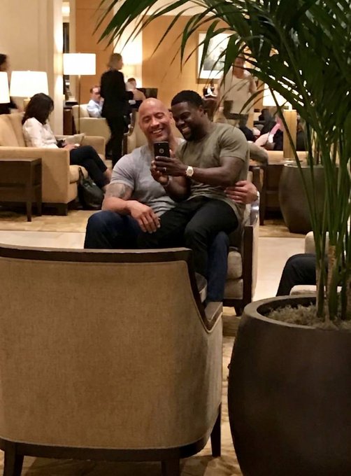 The image shows two men sitting together, one of them holding a cell phone while looking at something on it. They are both smiling, indicating that they might be sharing some good news or an amusing moment captured by the man with the cell phone. The setting appears to be a hotel lobby, as there is a potted plant in the background and several other people present in the scene.