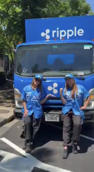 Two women wearing blue Ripple jerseys pose in front of a blue Ripple-branded truck with a license plate reading "134026," visually representing Ripple's on-the-ground promotional efforts to drive XRP adoption as the post highlights "THE ROAD TO XRP ADOPTION RUNS THROUGH THE CLARITY ACT."