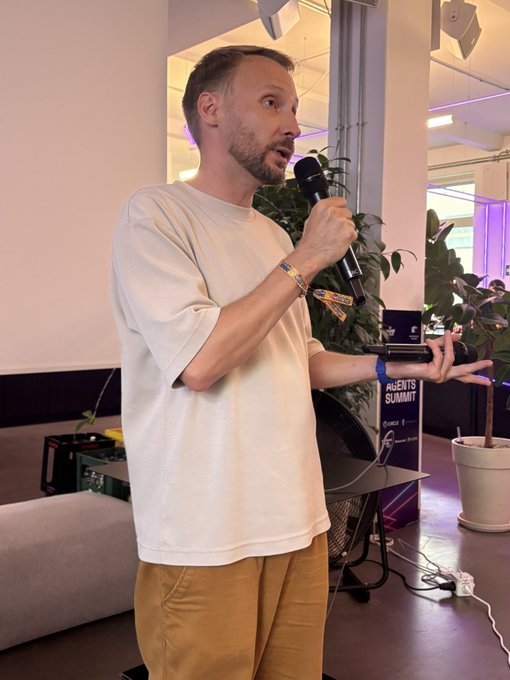The image shows a man standing up, holding a microphone, and speaking into it. He appears to be giving a speech or presentation. There are two potted plants visible in the scene - one near the left side of the frame and another on the right side. The background features a white wall with a window that allows natural light to enter the room. A sign is also present, which reads "Let the pitches begin".