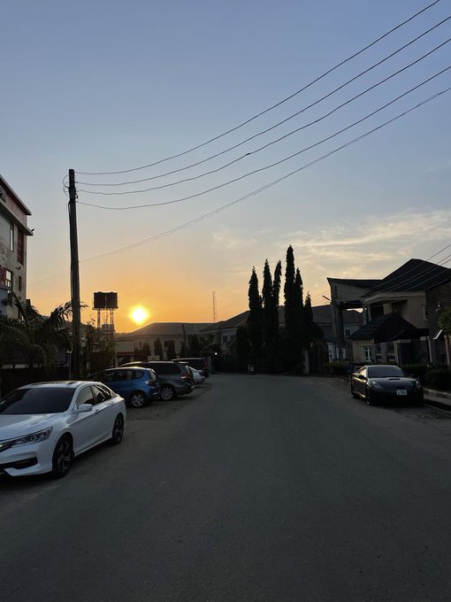 A suburban street at sunset with the sun low on the horizon, silhouetting trees, parked cars, and power lines against a warm orange and blue sky.
